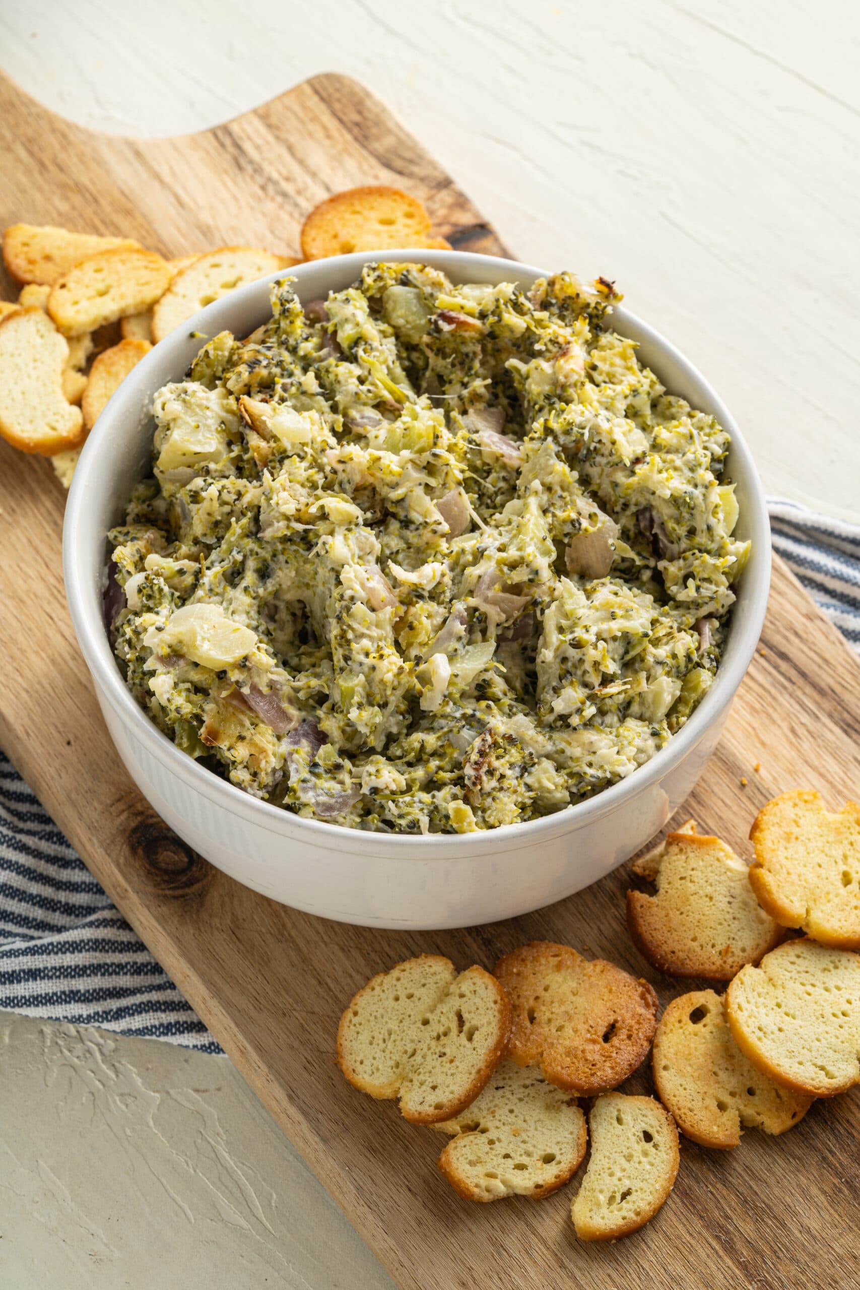 Picture of cooked broccoli cheese dip  in a white bowl, on a narrow rectangular tray with scattered crackers on both sides, which is on a blue and white striped towel.