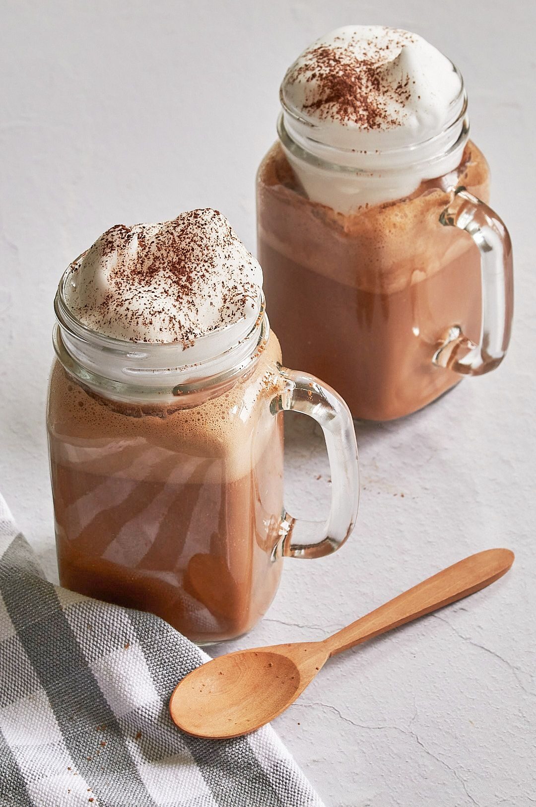 Two clear glass mason jar mugs with hot chocolate with whipped cream dripping on top with a wooden spoon in front of them and a grey and white checkered towel in bottom left corner.