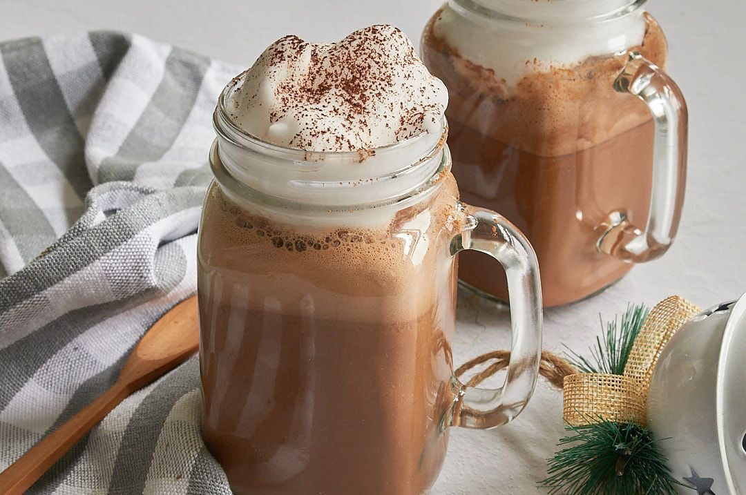 Horizontal picture of Two clear glass mason jar mugs with hot cocoa with whipped cream dripping on top with a wooden spoon on a grey and white striped towel in bottom left corner and a small piece of evergreen in the bottom right corner.