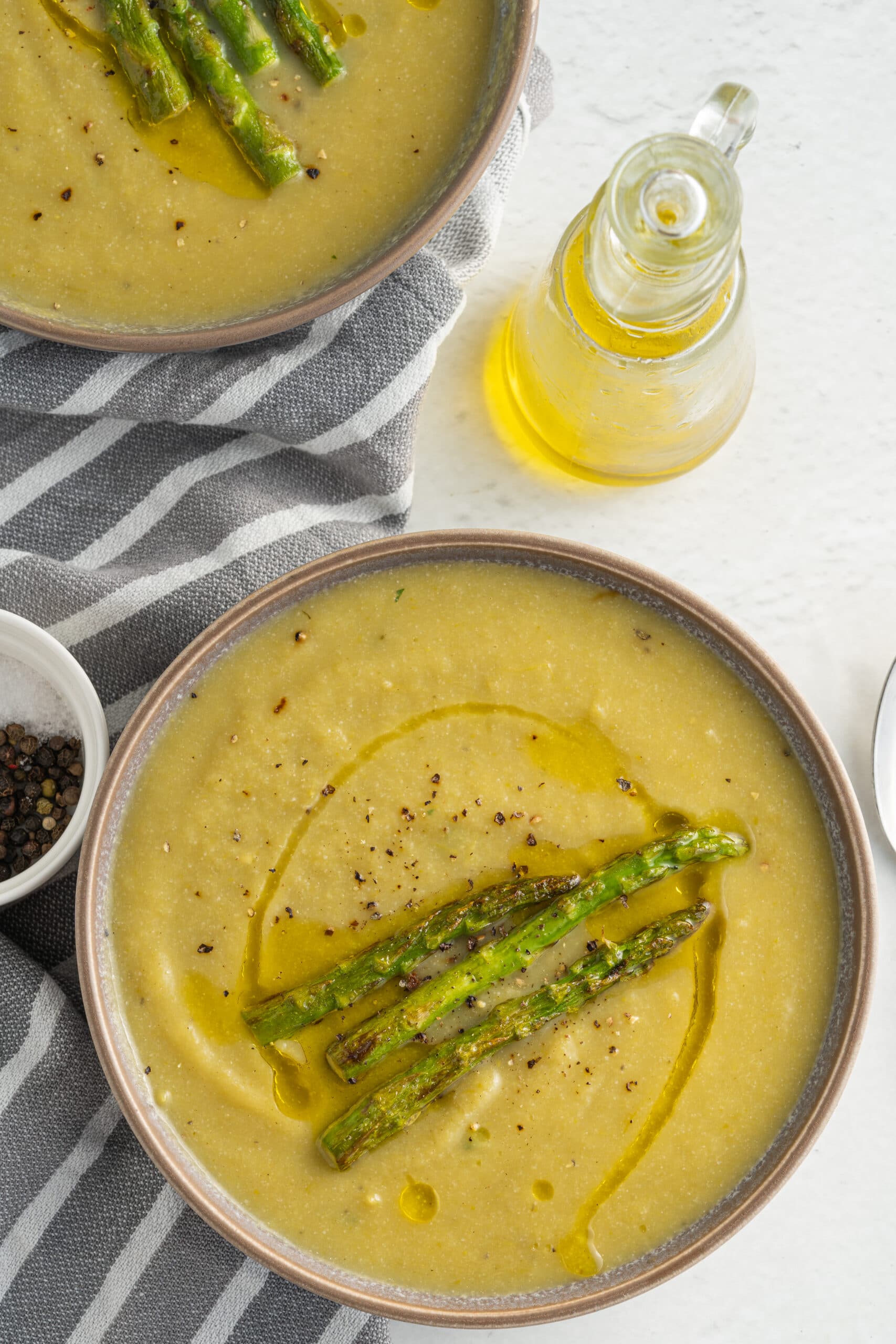 Creamy asparagus soup in a gray bowl with three asparagus spears on top and a bottle of olive oil behind it.