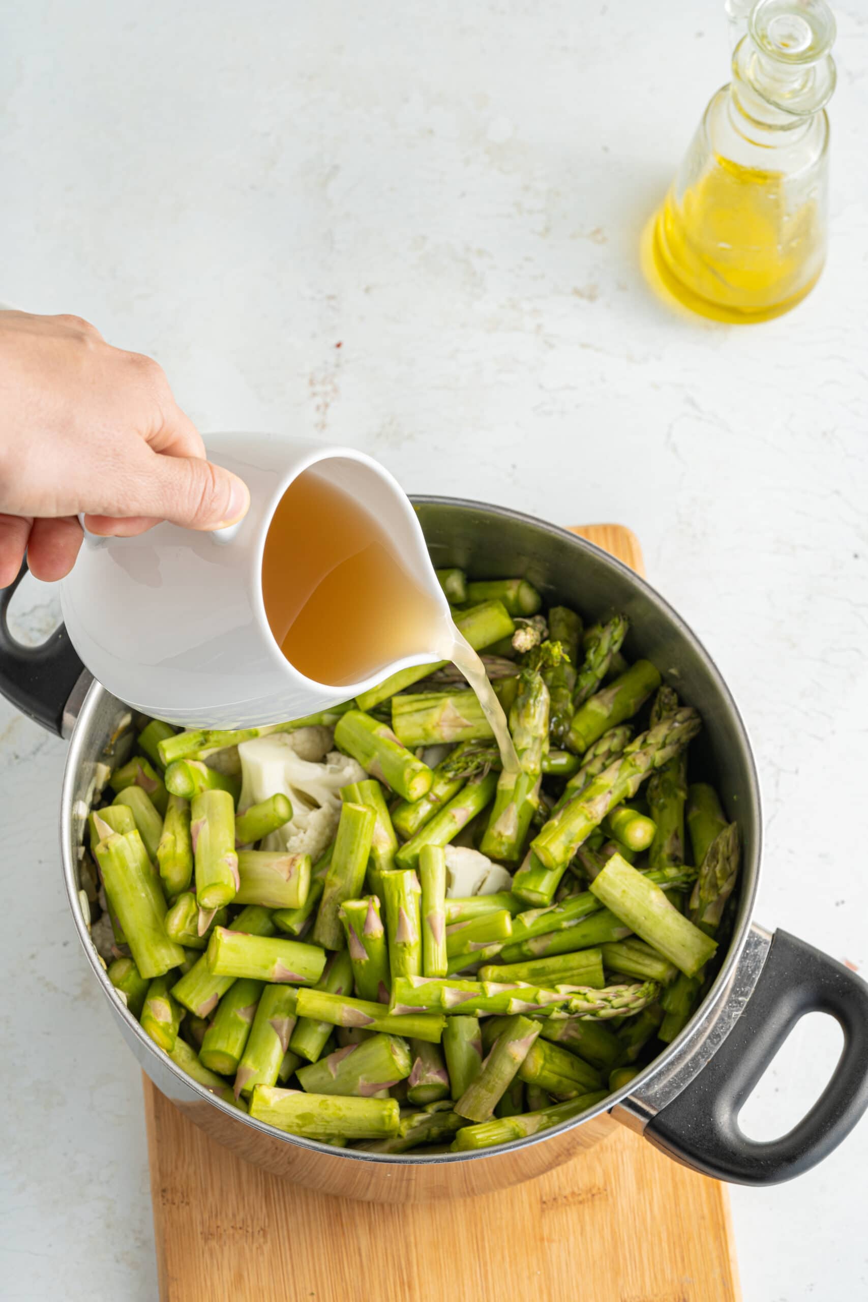 Chopped asparagus in a skillet with a white pitcher of broth about to be poured in the skillet.