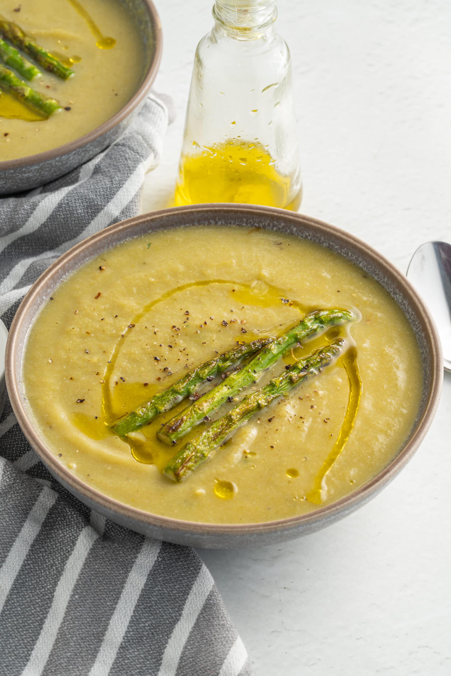 Creamy asparagus coup in a grey bowl with three asparagus spears in the middle and a grey and white striped towel to the left of it.