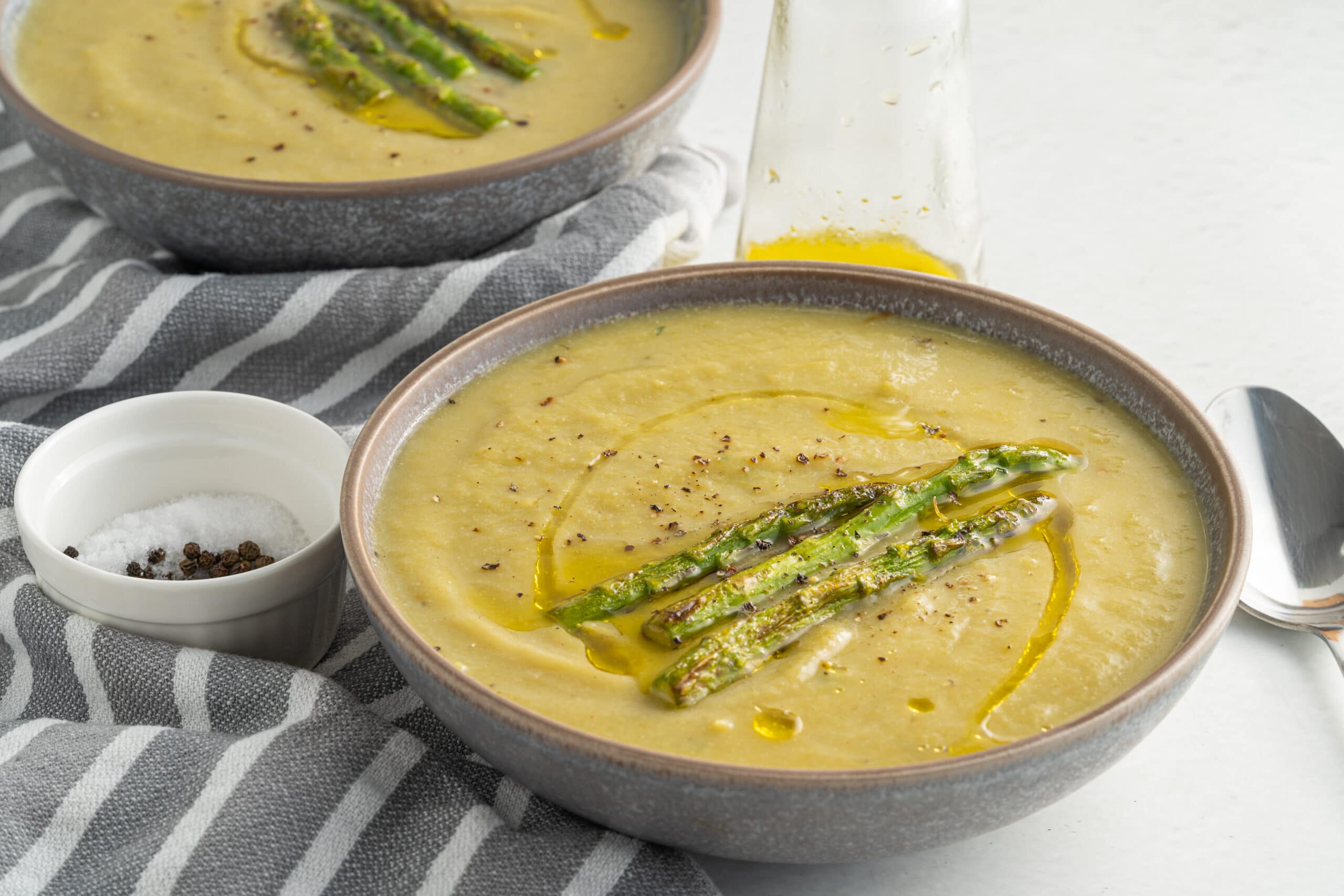 Creamy asparagus soup in a grey bowl with three asparagus spears in the middle and a small white bowl with salt and pepper in it.