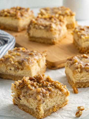 Picture of pecan cheesecake bars on a white cloth and others on a wood board in the background.
