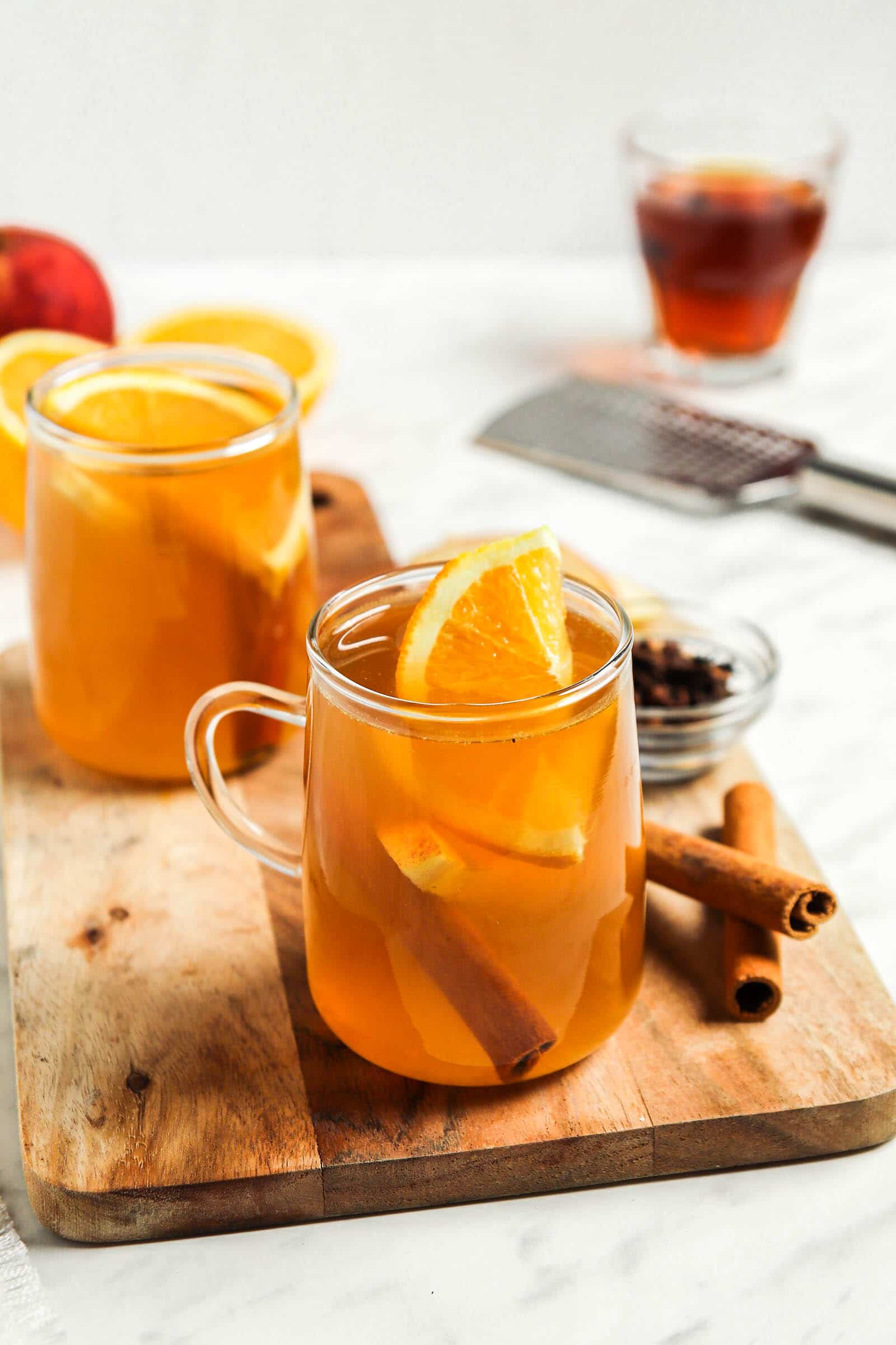 Two clear glasses of spiked apple cider on a wood board with sliced orange in them.
