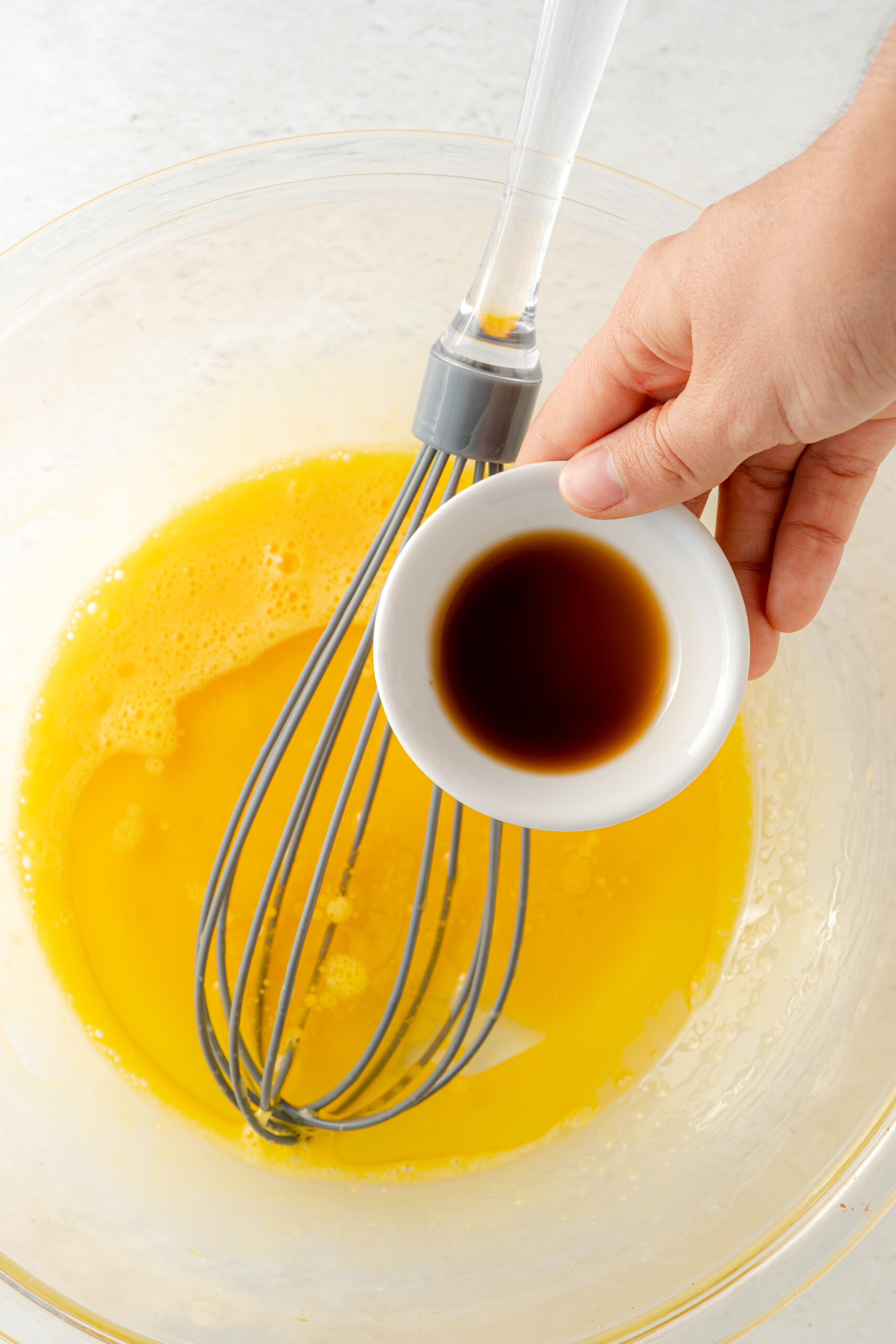 Eggs in large mixing bowl with whisk and small white bowl of vanilla being added in.