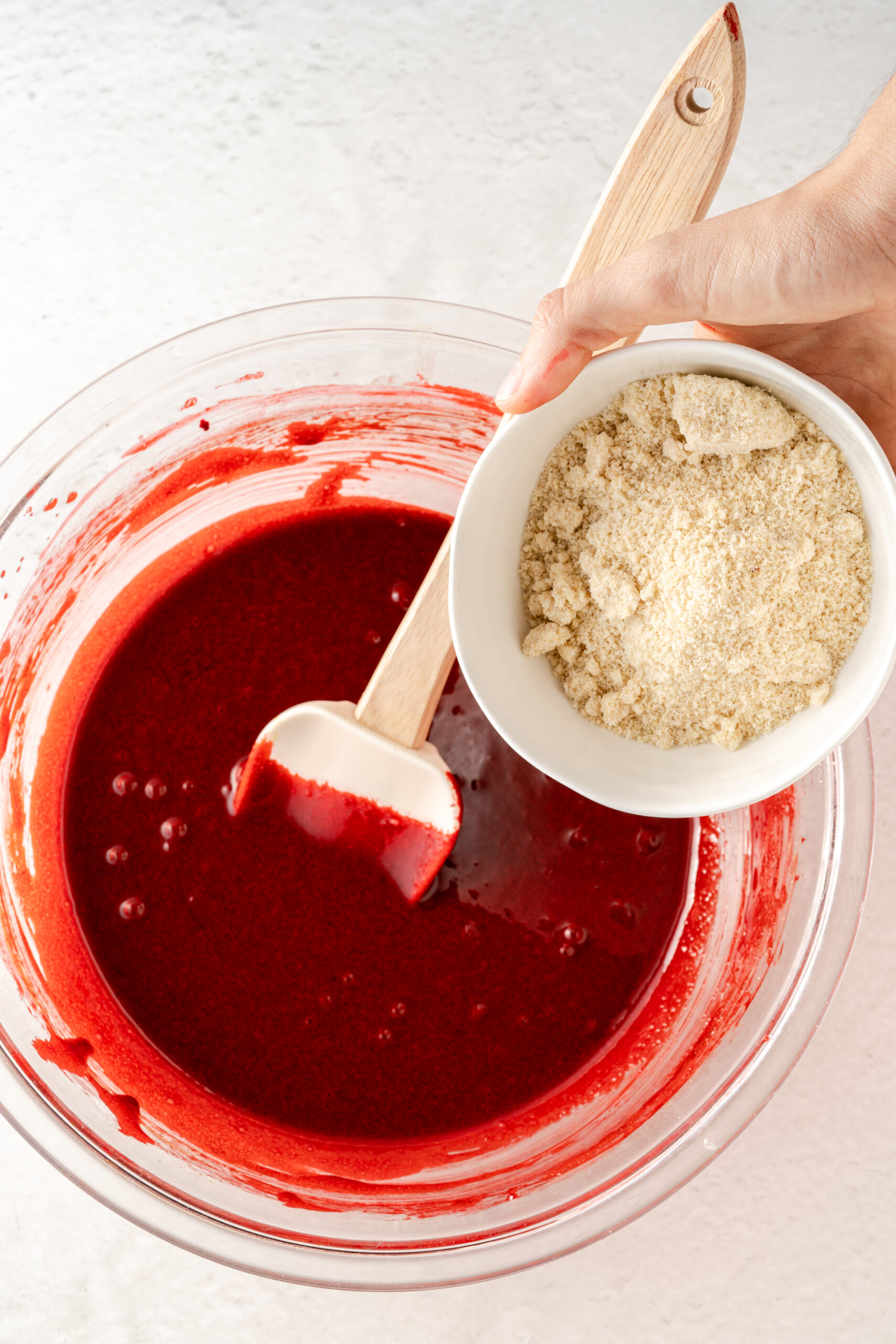 Red mixture in large mixing bowl with whisk and small white bowl of almond flour being added in.