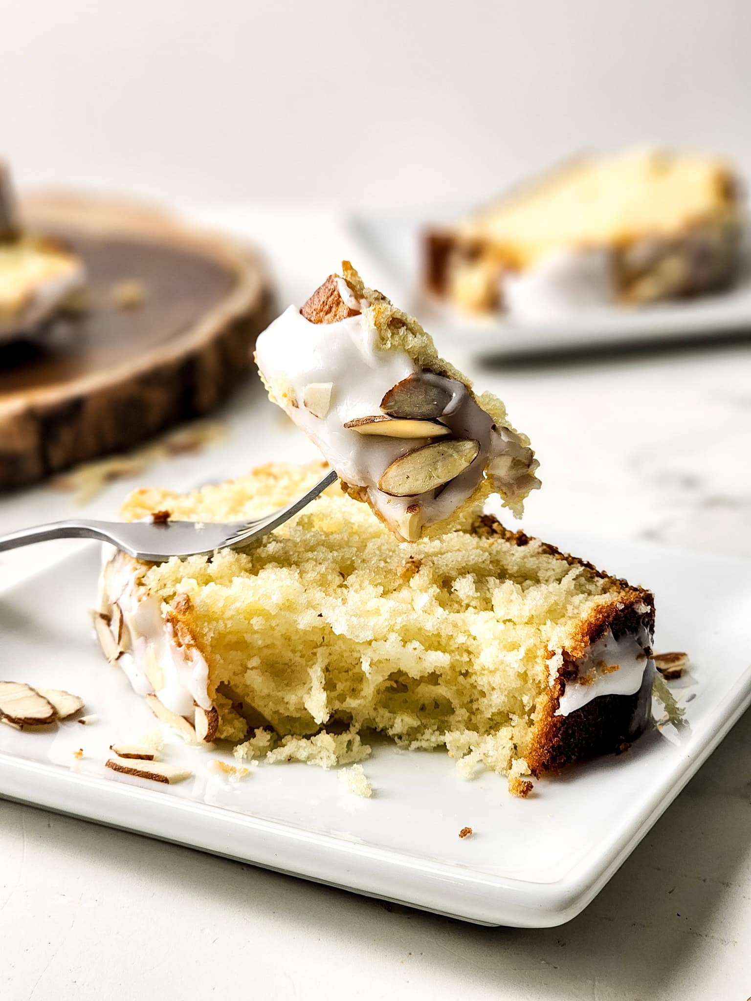 A slice of moist Gluten Free Greek Yogurt Almond Cake topped with a light almond glaze and sliced almonds, served on a white plate with a fork holding a bite of cake above the slice. Additional slices of almond cake are softly blurred in the background.