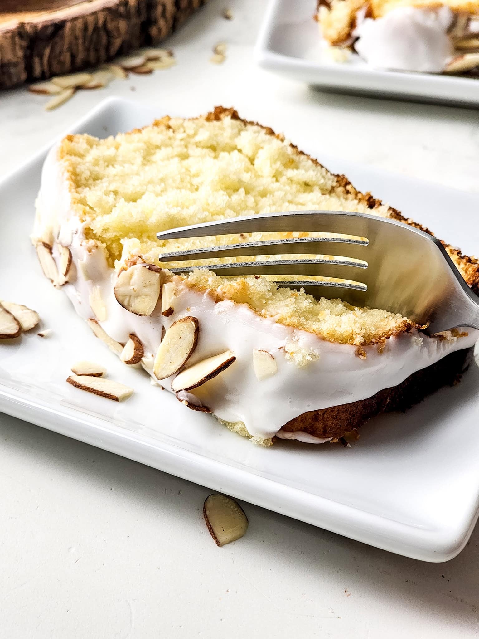 A slice of moist Gluten Free Greek Yogurt Almond Cake topped with a light almond glaze and sliced almonds, served on a white plate with a fork in the middle of the slice of cake. 