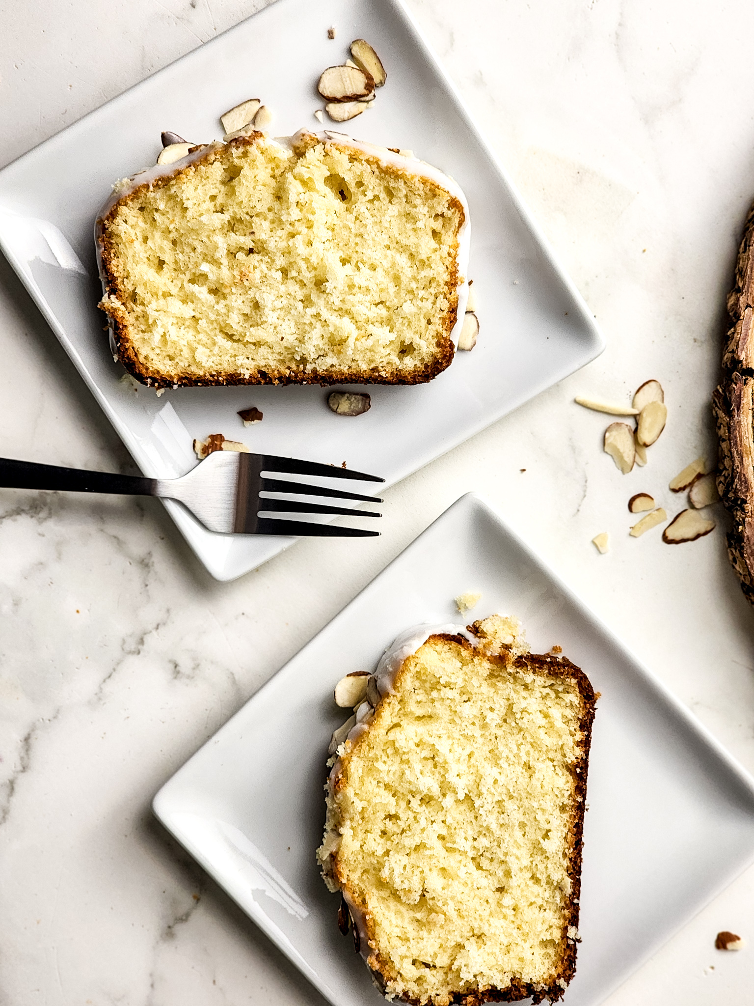 A slice of moist Gluten Free Greek Overhead picture of Yogurt Almond Cake topped with a light almond glaze and sliced almonds, served on a white plate with a fork next to the slice of cake. 