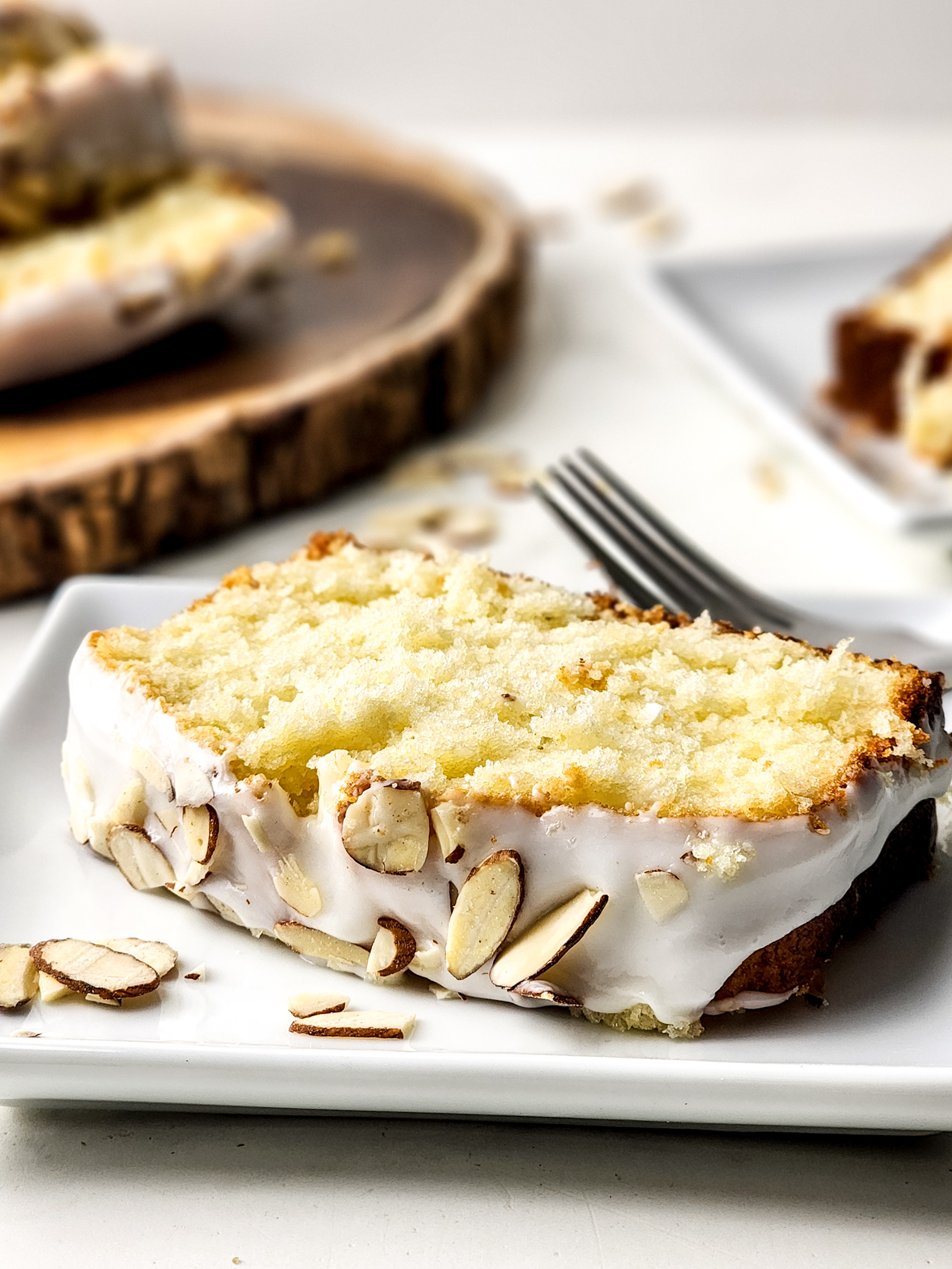 A slice of gluten free Greek yogurt almond cake topped with light almond glaze and sliced almonds on a white plate.