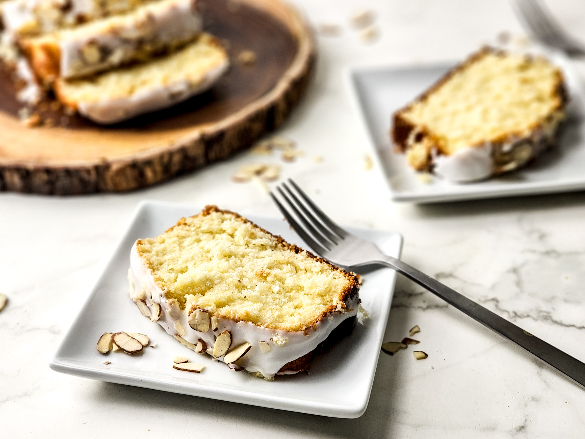 A slice of moist Gluten Free Greek Yogurt Almond Cake topped with a light almond glaze and sliced almonds, served on a white plate with a fork next to the slice of cake. Another slice is on another white plate behind it and there is a wodd tray with ore slice of cake on the other side.