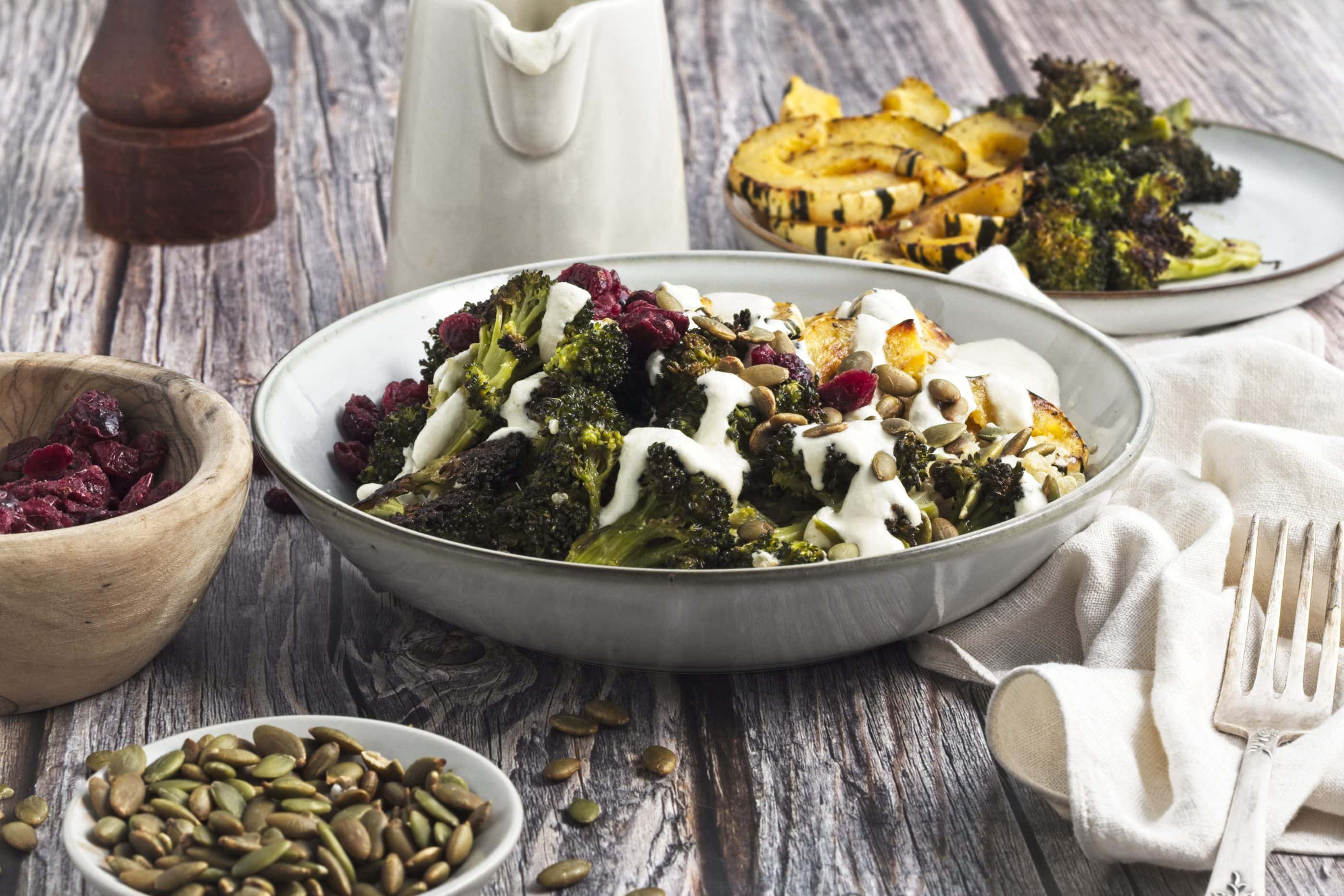 Bowl of Roasted Broccoli Delicata Squash Quinoa Bowl with dressing and pumpkin seeds on top next to a small bowl of pumpkin seeds and a plate of roasted broccoli ad squash above it.