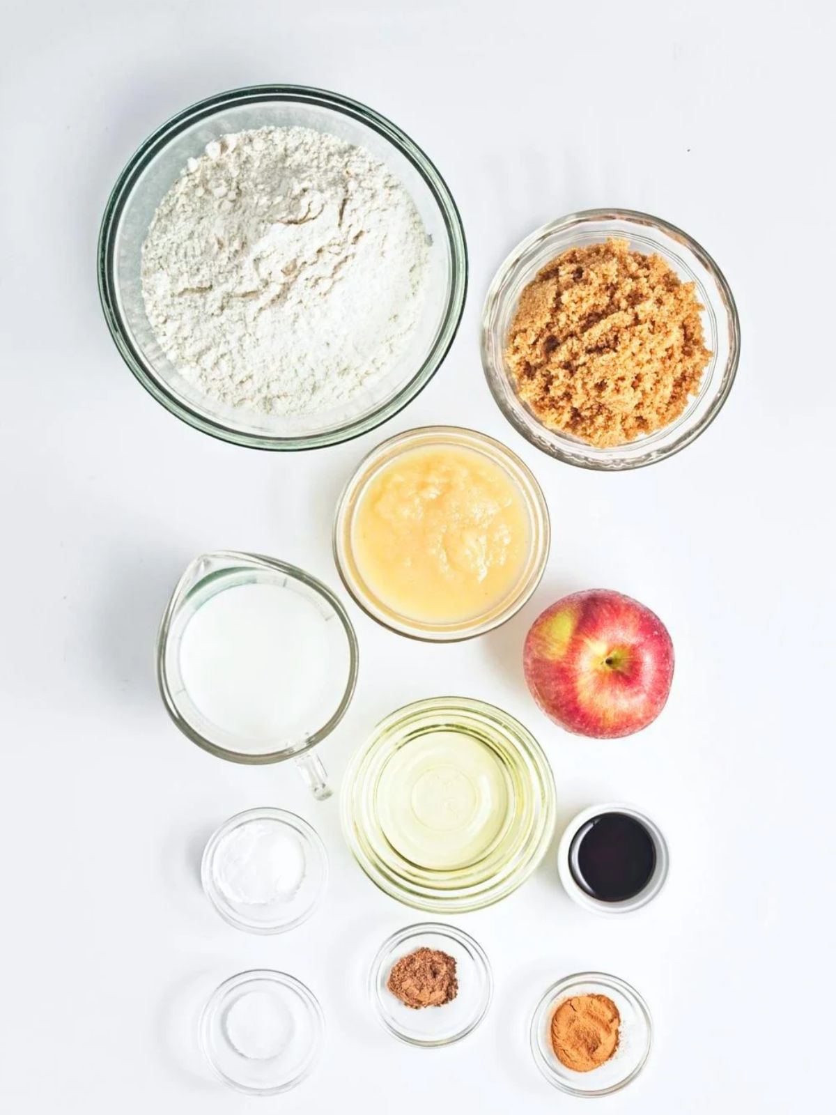 Ingredients for vegan apple crumb muffins arranged in glass bowls, including flour, brown sugar, applesauce, plant-based milk, chopped apple, oil, vanilla extract, baking powder, baking soda, and cinnamon.
