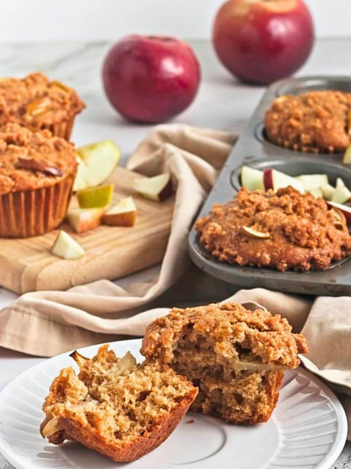 A vegan apple crumb muffin cut in half to show its soft, moist interior with apple pieces and a golden cinnamon crumb topping, served on a white plate with more muffins in the background.