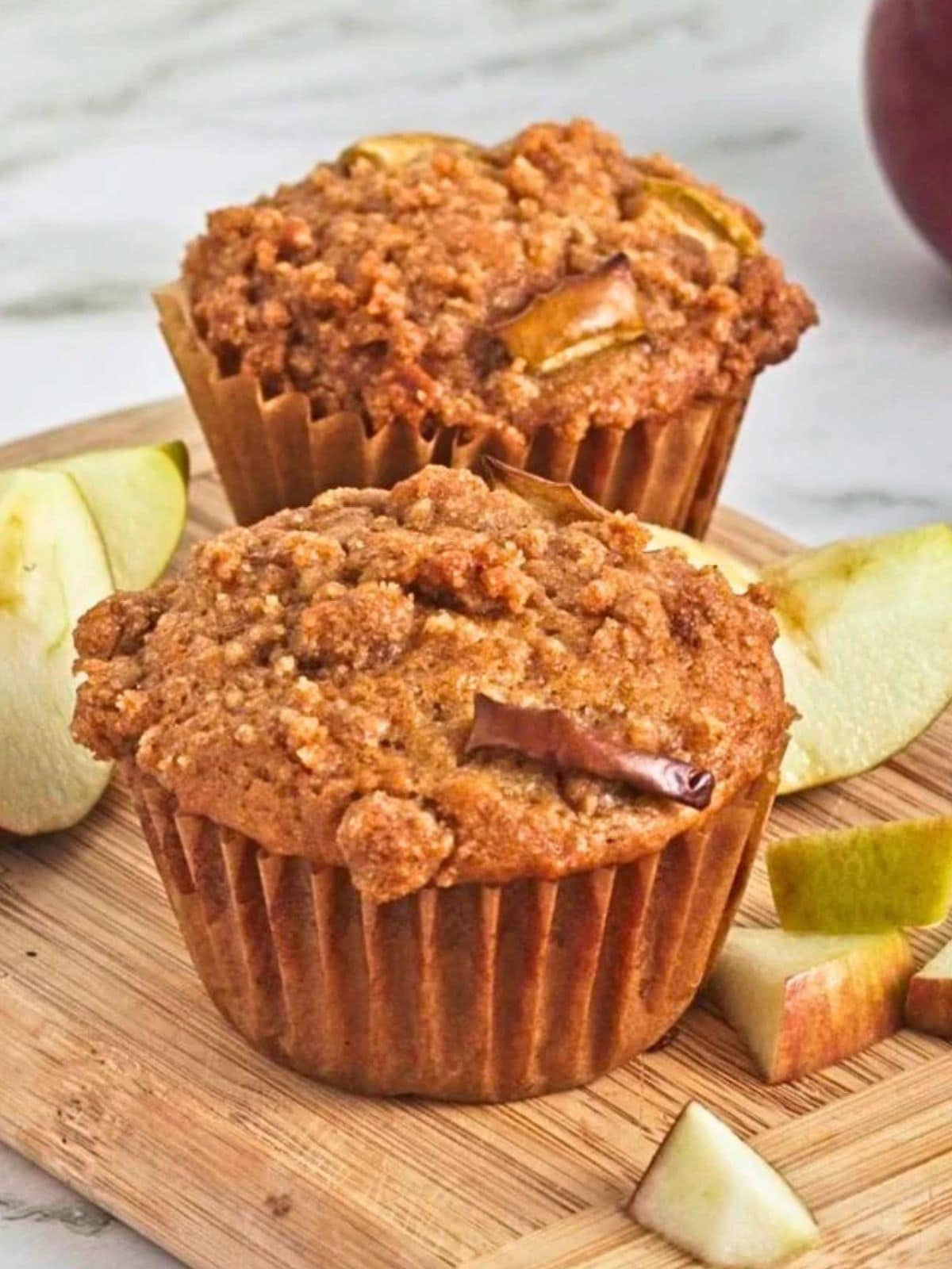 Two homemade apple crumb muffins with a crunchy cinnamon streusel topping on a wooden cutting board, surrounded by fresh apple slices.
