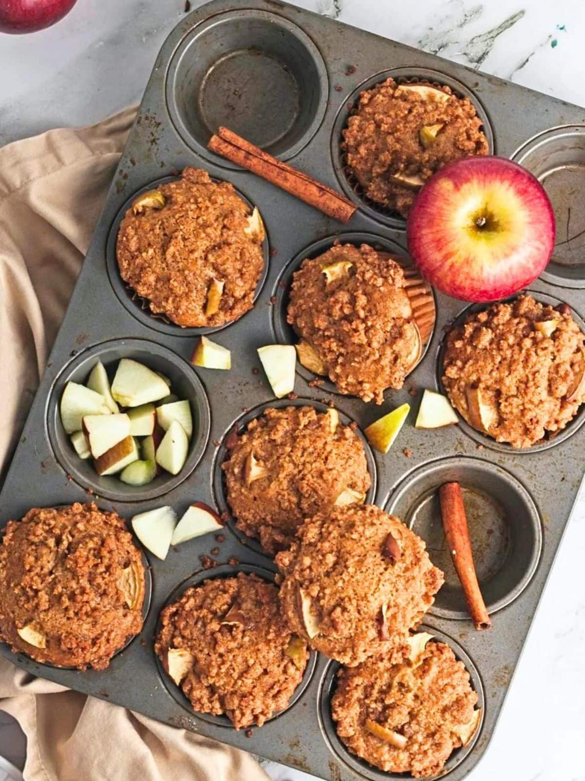 Overhead view of vegan apple crumb muffins with cinnamon crumb topping in a metal muffin pan, surrounded by fresh apple pieces and cinnamon sticks.