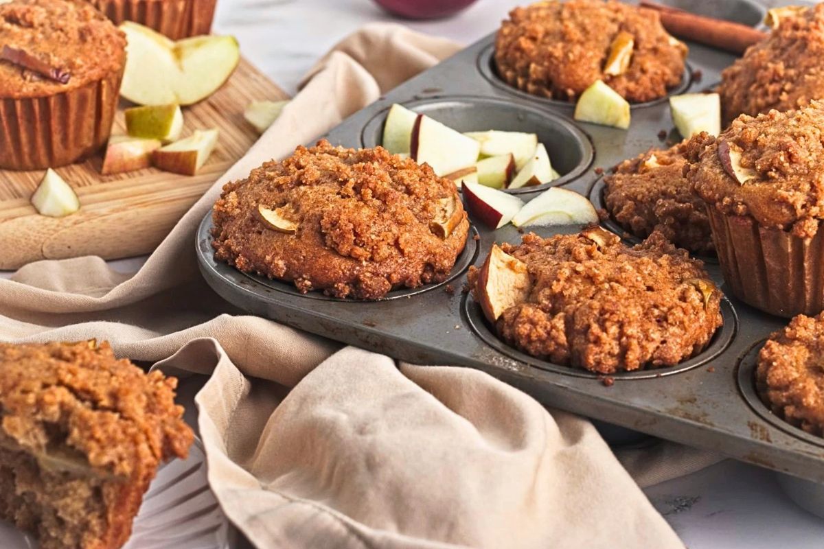 Freshly baked apple crumb muffins with a golden cinnamon streusel topping in a muffin tin, with sliced apples, whole apples, and a beige kitchen towel arranged around them.