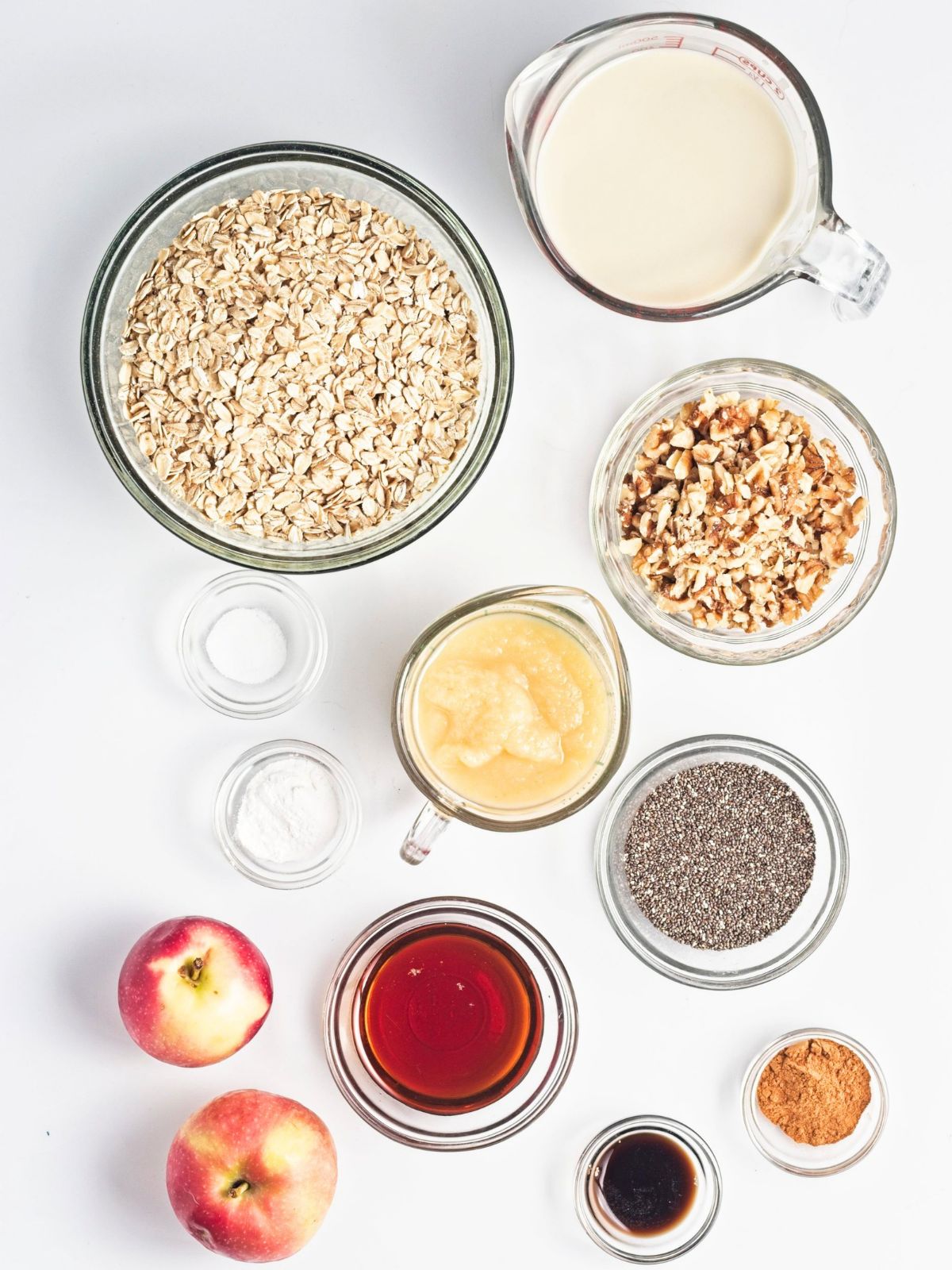 Ingredients for vegan baked apple protein oatmeal arranged on a white surface, including rolled oats, apples, walnuts, chia seeds, applesauce, plant-based milk, cinnamon, maple syrup, and baking ingredients.