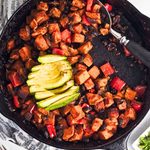 Sweet potato black bean hash cooked in a cast iron skillet with roasted sweet potatoes, black beans, red bell peppers, and red onion, topped with sliced avocado.