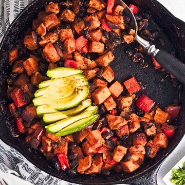 Sweet potato black bean hash cooked in a cast iron skillet with roasted sweet potatoes, black beans, red bell peppers, and red onion, topped with sliced avocado.