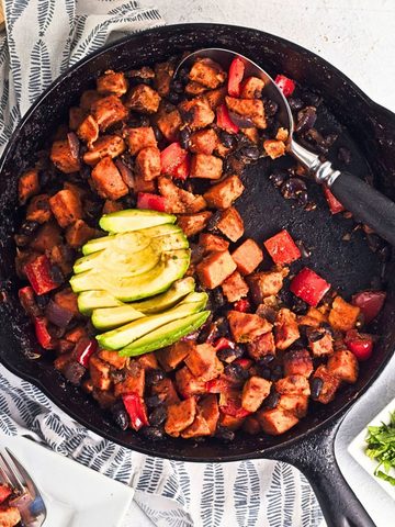 Sweet potato black bean hash cooked in a cast iron skillet with roasted sweet potatoes, black beans, red bell peppers, and red onion, topped with sliced avocado.