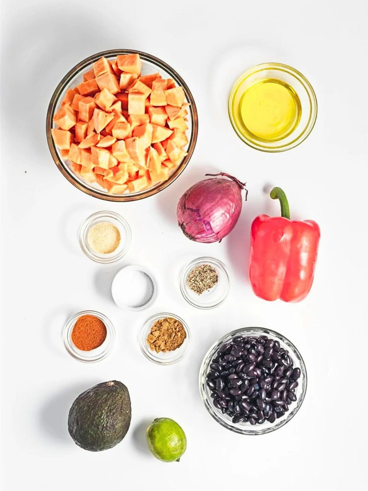 Ingredients arranged on a white surface, in individual glass bowls, except the onion, red pepper, avocado and lime which are around the bowls.