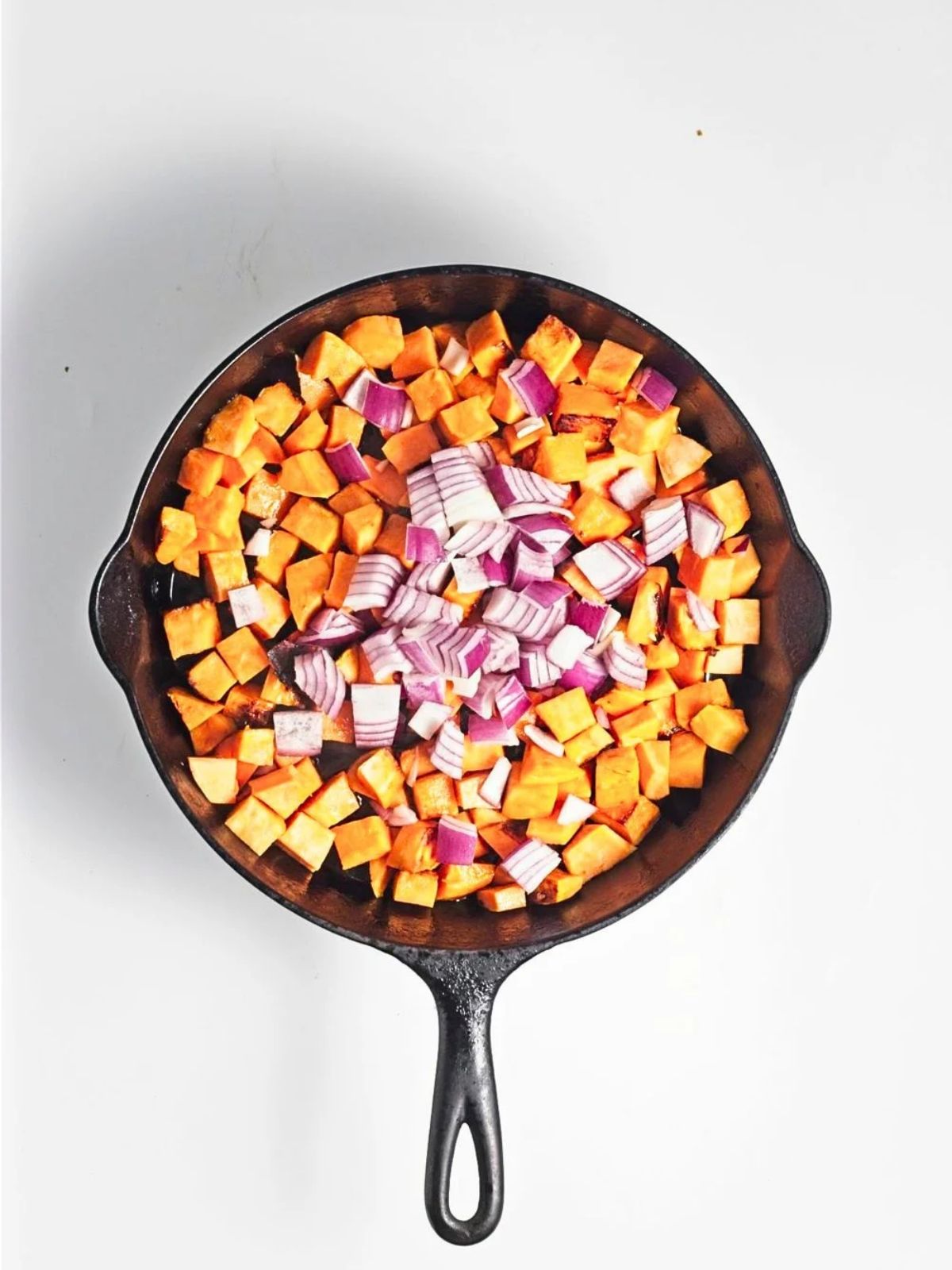 Diced sweet potatoes cooking in a cast iron skillet with chopped red onion added.
