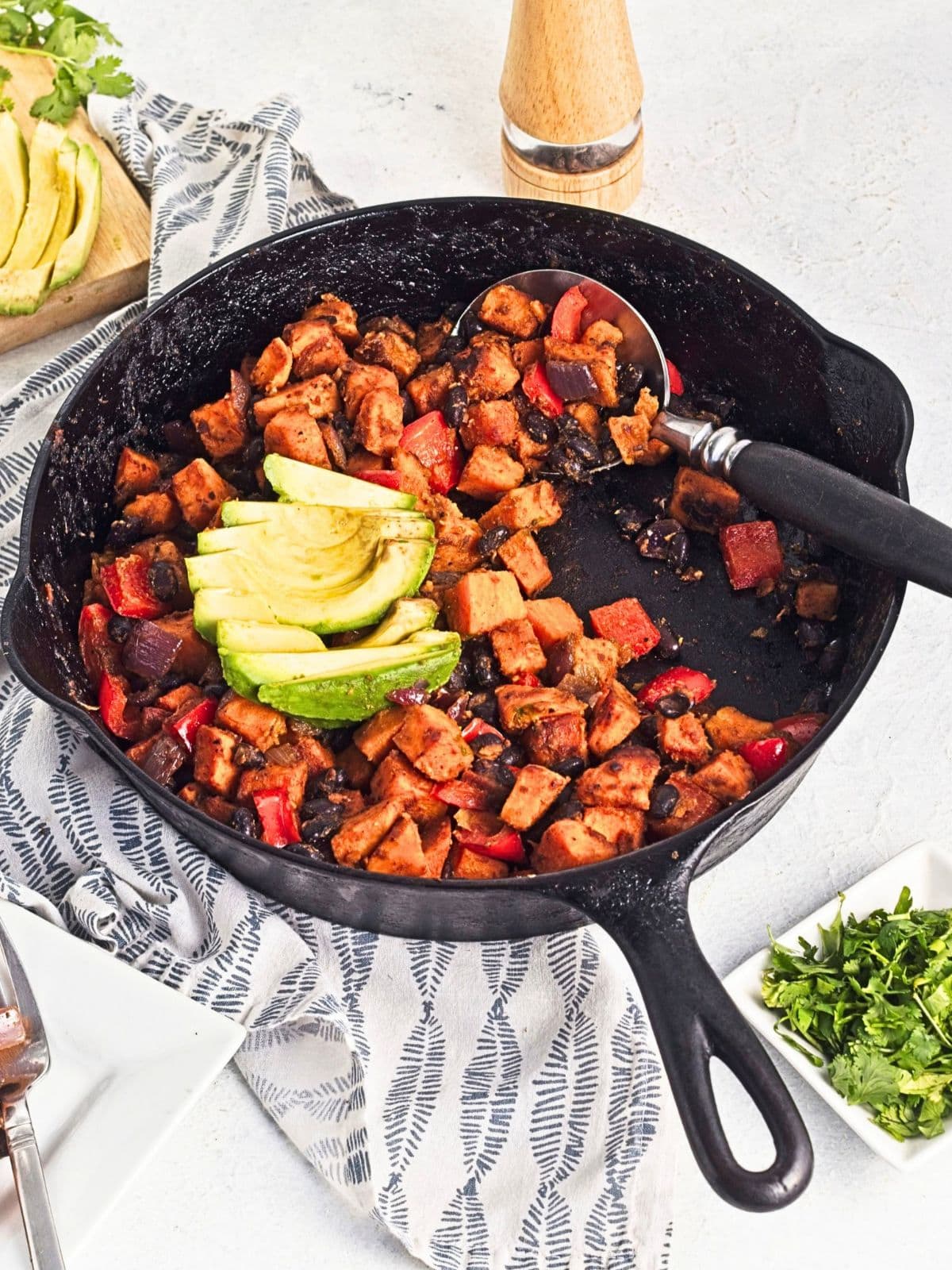 Sweet potato black bean hash cooked in a cast iron skillet with roasted sweet potatoes, black beans, red bell peppers, and red onion, topped with sliced avocado with a spoon in the skillet.
