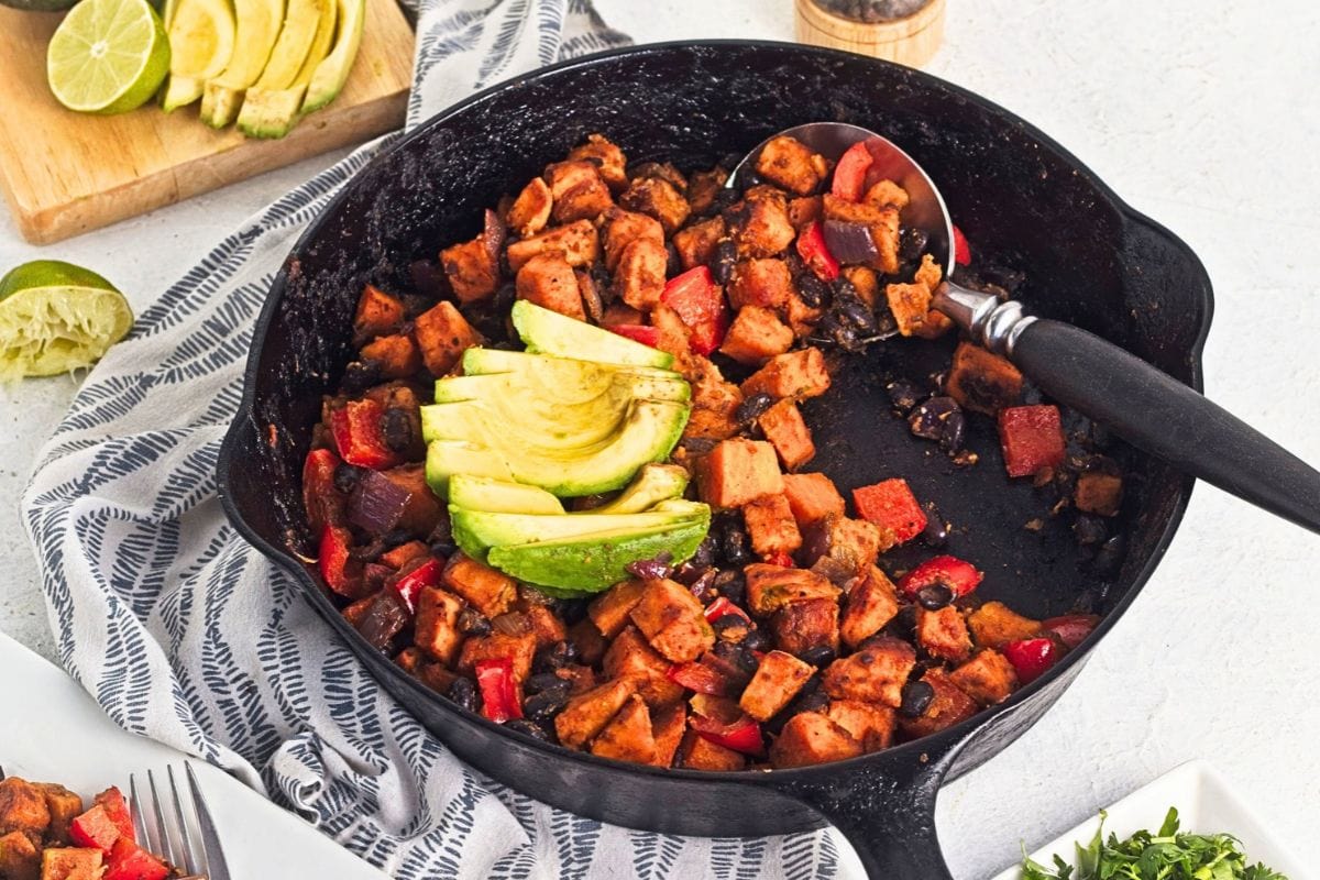 Cooked hash cooked in a dark cast iron skillet with a spoon, topped with sliced avocado.